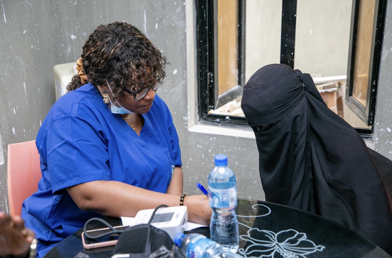 A MED-WECARE health worker in blue scrubs records notes beside a blood pressure monitor during a consultation with a community member.