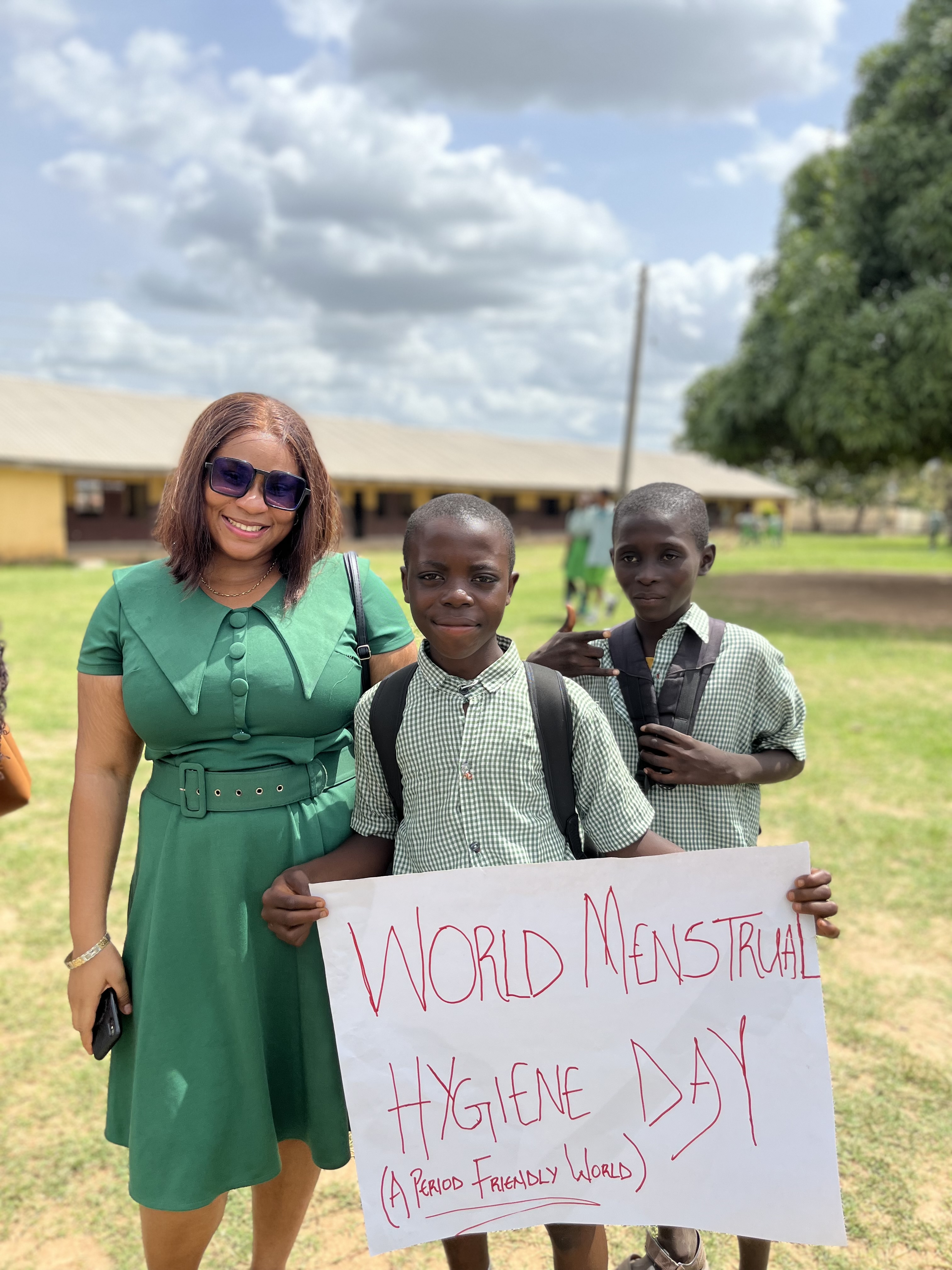 A MED-WECARE team member stands with two schoolboys, one holding a sign that reads 'World Menstrual Hygiene Day — A period friendly world'.